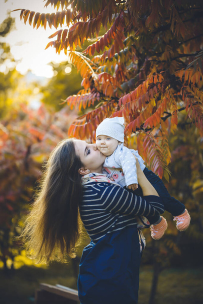 Όμορφη γυναίκα σε ένα φόρεμα παίζει με ένα παιδί στην αγκαλιά της, στέκεται στην φθινοπωρινή φύση με πεσμένα φύλλα. Νεαρό κορίτσι με μακριά μαλλιά χαλαρώστε στο πάρκο με κόκκινα κίτρινα φύλλα. Ευτυχισμένη οικογένεια. - Φωτογραφία, εικόνα