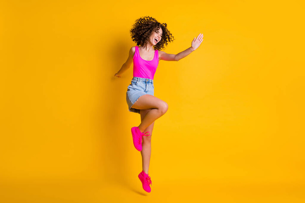 Full body photo of attractive curly lady jump up high dancing students party delighted emotions wear pink tank top denim mini skirt shoes isolated vivid bright yellow color background - Foto, immagini