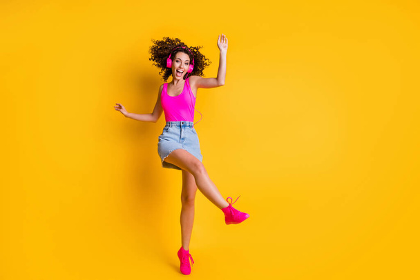 Full body photo of crazy charming curly lady dance carefree raise leg student party delighted listen earphones wear pink tank top denim mini skirt shoes isolated bright yellow color background - Foto, Imagem