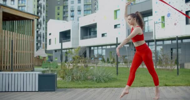 Mujer atlética joven en rojo realiza voltereta con cinta de gimnasia en el patio de un edificio de apartamentos, gimnasia en la ciudad, entrenamiento al aire libre, deportes en cámara lenta, 4k 120p HQ Prores 10 bit - Imágenes, Vídeo