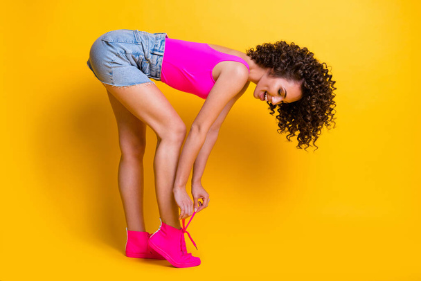 Full length profile photo of funny charming cute lady prepare summer chill event bend spine check shoelaces wear pink singlet denim skirt sneakers isolated shine yellow color background - Photo, Image