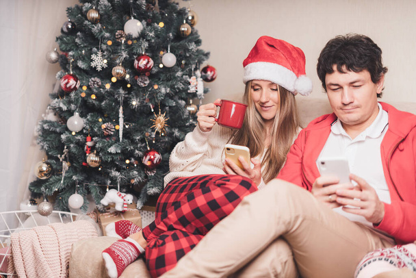 携帯電話と赤いサンタの帽子の美しい夫婦の夫と妻の肖像画自宅でクリスマスの冬の休日を楽しむ. - 写真・画像