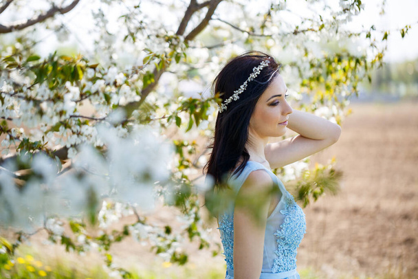 Όμορφο κορίτσι σε ένα ελαφρύ καλοκαιρινό μπλε μακρύ φόρεμα στολισμένο στα μαλλιά της ενάντια σε ένα ανθισμένο δέντρο. Τρυφερή προσωπογραφία μιας νεαρής γυναίκας με λευκά άνθη - Φωτογραφία, εικόνα