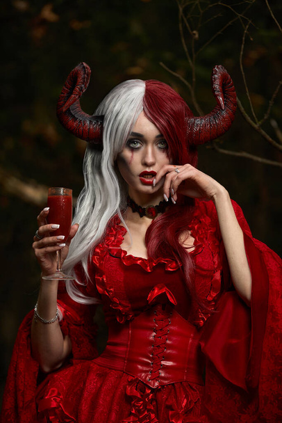 Maleficent Woman in Red Clothing and Horns in dark Forest. Posing with wineglass. - Photo, Image