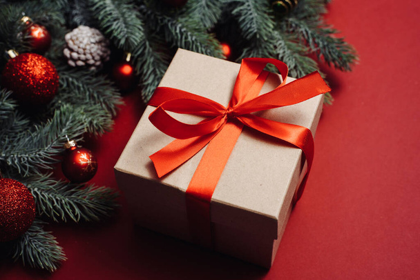 Χειροποίητο δώρο με κόκκινη σατέν κορδέλα δίπλα σε κλαδιά και μπάλες χριστουγεννιάτικου δέντρου, παιχνίδια σε κόκκινο φόντο - Φωτογραφία, εικόνα