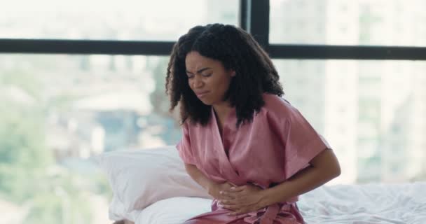 胃の痛みに苦しんでいる病気の黒人女性は、ベッドに座って、午前中に腹部や月経痛を感じて - 映像、動画
