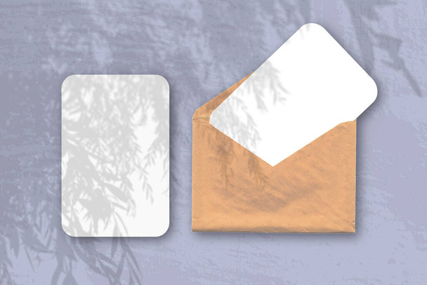 Ein Umschlag mit zwei Blättern strukturierten weißen Papiers auf dem blauen Hintergrund des Tisches. Mockup-Overlay mit den Schatten der Pflanze. Natürliches Licht wirft Schatten von einem Weidenast - Foto, Bild