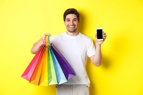 Joven sosteniendo bolsas de compras y mostrando la pantalla del teléfono móvil, aplicación de dinero, de pie sobre el fondo amarillo - Foto, Imagen