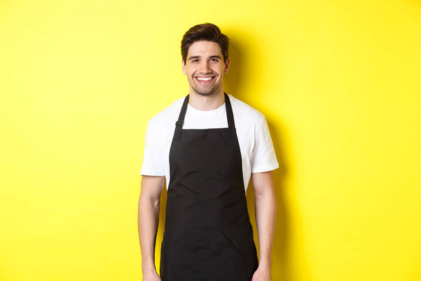 Camarero joven en delantal negro, sonriente, trabajando en tienda o cafetería, de pie sobre fondo amarillo - Foto, imagen