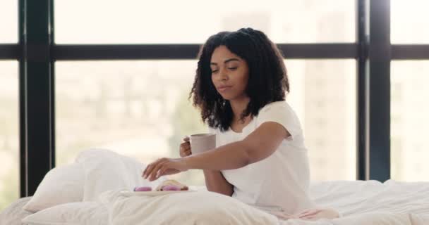 Giovane signora afroamericana godendo la colazione a letto, mangiare biscotti con caffè al mattino - Filmati, video