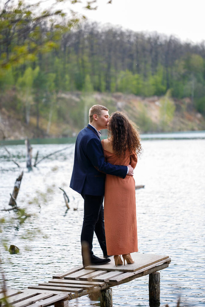 A young couple holding hands on a wooden bridge in the middle of a blue lake. Masonry on the island on a background of trees. Nature, landscape. Romance and love, happy couple - Photo, Image