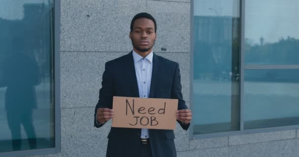 Ritratto all'aperto di giovane impiegato afroamericano in piedi vicino a un edificio per uffici con poster NEED JOB - Filmati, video