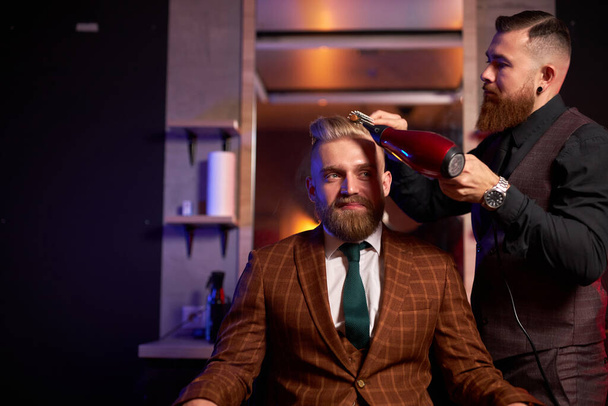 stylish man sitting in barber shop while hairdresser dry his hair - Фото, зображення