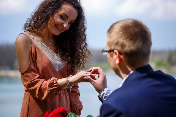 Lake in the forest: a man made a surprise to his girlfriend, he puts a ring on her finger, a declaration of love and an offer of marriage. Girl with a smile on her face and a bouquet of red roses - Foto, Imagem