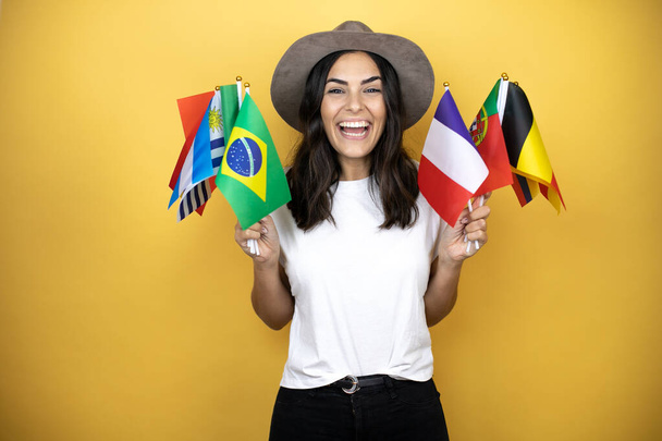 Beautiful woman wearing casual white t-shirt and a hat standing over yellow background holding many country flags - Photo, Image