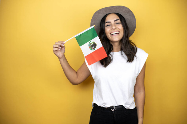 Beautiful woman wearing casual white t-shirt and a hat standing over yellow background holding the mexican flag - Photo, Image
