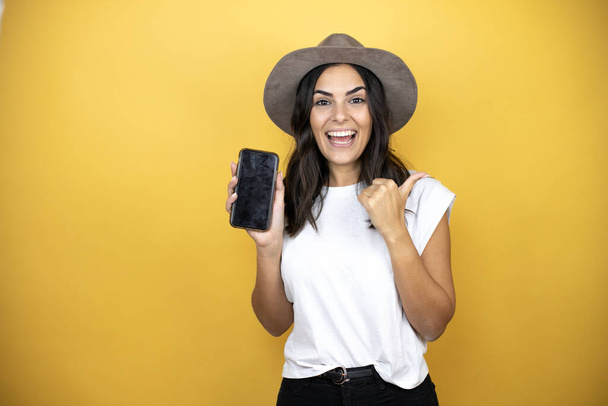 Beautiful woman wearing casual white t-shirt and a hat standing over yellow background smiling and pointing to the copy space - Foto, immagini