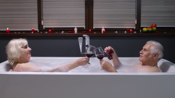 Casal sênior avô e avó está tomando banho espumoso, bebendo vinho tinto no banheiro - Filmagem, Vídeo