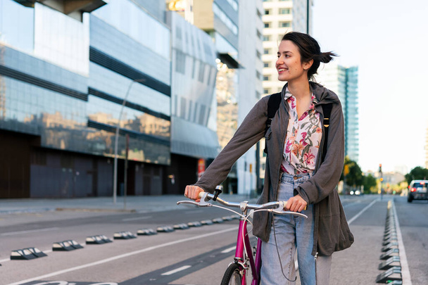 happy young girl smiling and walking with a pink retro bicycle by the bike path at city, concept of active lifestyle and sustainable mobility - Zdjęcie, obraz