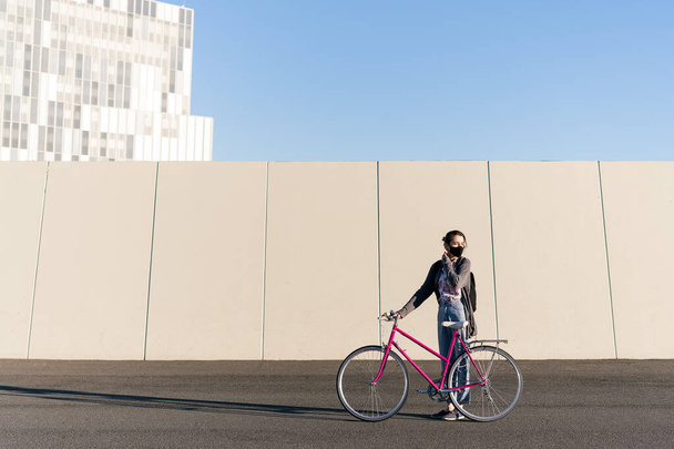 young girl with a pink retro bike at city adjusting her protective mask, concept of active lifestyle, protection against covid and sustainable urban mobility - Photo, image