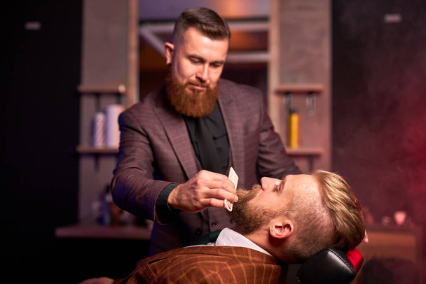young caucasian handsome man on chair of barber having procedure of shaving - Zdjęcie, obraz