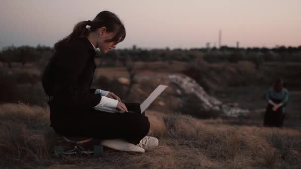 Câmera gira em torno de menina de estilo moderno trabalhando em laptop em um campo no pôr do sol - Filmagem, Vídeo