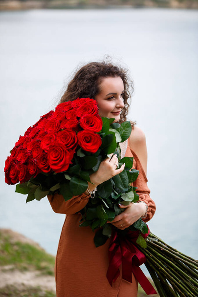Ein schönes Mädchen von europäischem Aussehen mit lockigem Haar und einem Lächeln auf dem Gesicht mit einem riesigen Strauß roter Rosen vor blauem See. Warmer Sommertag, glückliche junge Frau, Emotionen der Freude - Foto, Bild