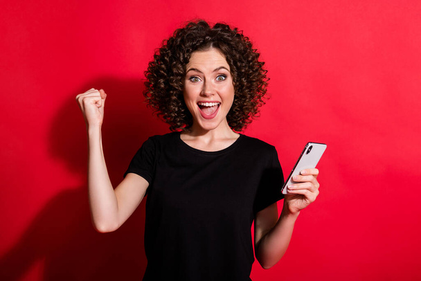 Fotografie štěstí vlnitý vítěz dáma oblečený ležérní černá outfit držení telefonu rostoucí pěst izolované červené barvy pozadí - Fotografie, Obrázek