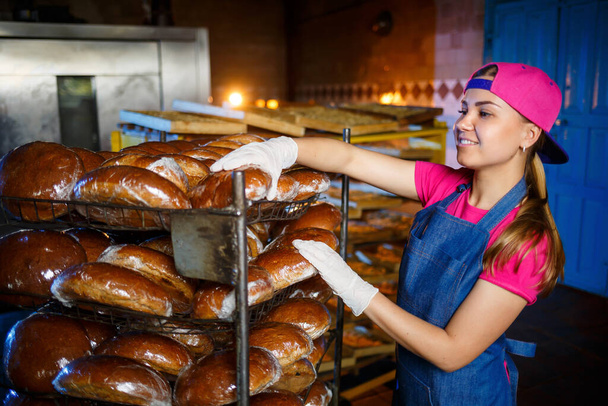 Professional baker - a young, pretty woman in a jeans apron holds fresh bread against the background of a bakery or bakery. Bakery products. Bread production - Photo, Image