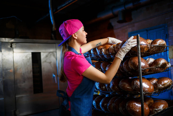 Ein junges Mädchen arbeitet in einer Bäckerei. Sie stellt Brot in ein Regal. Bäckerin am Arbeitsplatz in einer Bäckerei. Ein professioneller Bäcker hält Brot in den Händen. Brotproduktionskonzept - Foto, Bild