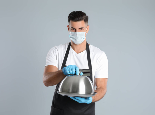 Waiter in medical face mask holding tray with lid on light grey background - 写真・画像