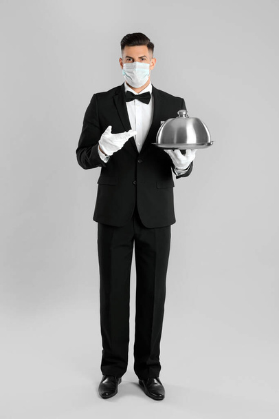 Waiter in medical face mask holding tray on light grey background - Fotoğraf, Görsel