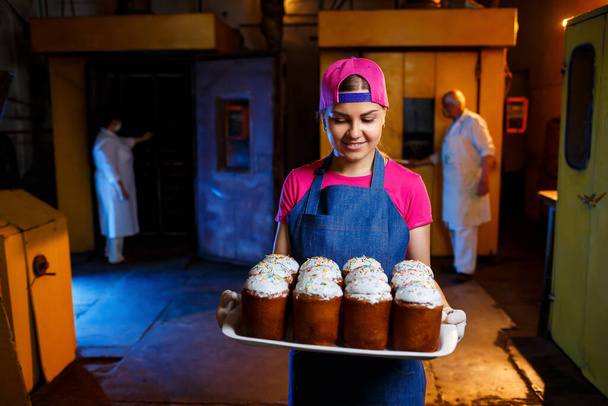 A péklány tálcát tart forró süteményekkel a pékségben. Farmerruhát és sapkát visel. Pékipari termékek gyártása. Rack friss ropogós süteményekkel. - Fotó, kép