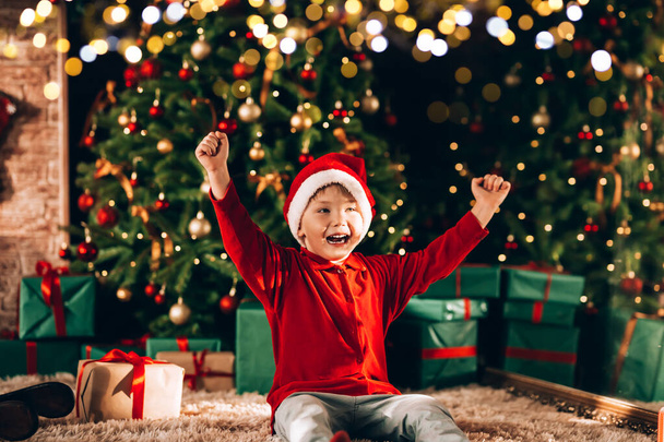 The boy has a santa hat on his head. In the background there is a large decorated Christmas tree with gifts in green packaging and a bow. Christmas mood. - Photo, Image