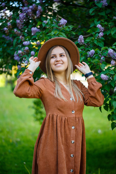 Portrait of a stylish girl in a brown hat and dress on a background of lilacs tree on a sunny warm day. A young woman of European appearance with a smile on her face - 写真・画像