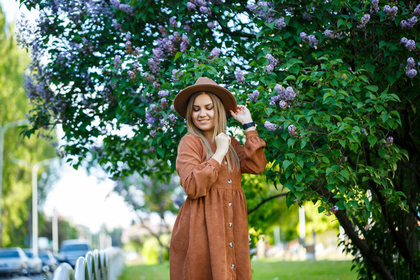Портрет стильной девушки в коричневой шляпе и платье на фоне сирени дерева в солнечный теплый день. Молодая женщина европейского образца с улыбкой на лице - Фото, изображение