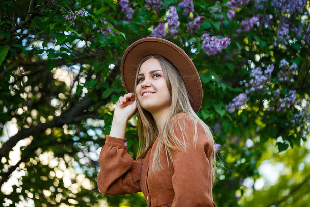 Симпатичный женский портрет в коричневой большой шляпе и стильная одежда на фоне сирени в ботаническом саду. Длинные светлые каштановые волосы лежат на плечах - Фото, изображение