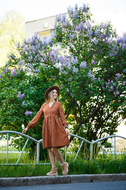 Cute girl portrait in brown big hat and stylish clothes against the background of a lilac tree in a botanical garden. Long light brown hair lies on the shoulders - Foto, Imagen