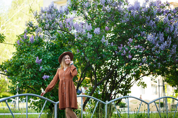 Симпатичный женский портрет в коричневой большой шляпе и стильная одежда на фоне сирени в ботаническом саду. Длинные светлые каштановые волосы лежат на плечах - Фото, изображение