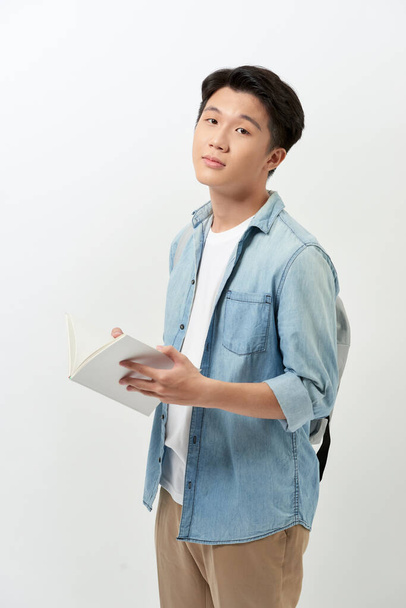 Joven hombre asiático leyendo libro y usando ropa casual con mochila sobre fondo blanco. - Foto, Imagen