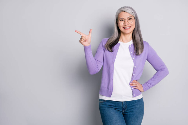 Photo portrait of positive optimistic grandmother smiling pointing showing at side with finger wearing jeans violet cardigan spectacles isolated on grey color background - Fotoğraf, Görsel