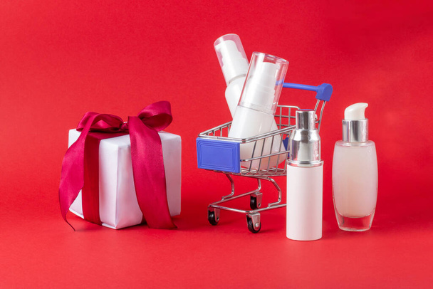 Set of face and body cosmetics, gift box and shopping trolley on a bright red background. The concept of buying cosmetics, online store, holiday - Photo, image