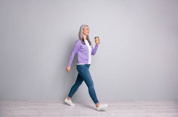 Full length body size photo of smiling elder woman having break going fast keeping cup of coffee wearing jeans cardigan eyeglasses isolated on grey color background - Fotografie, Obrázek