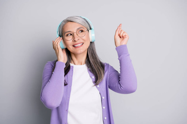 Photo portrait of dreamy smiling senior woman looking up pointing with finger listening to music touching wireless earphones wearing casual outfit isolated on grey color background - Fotografie, Obrázek