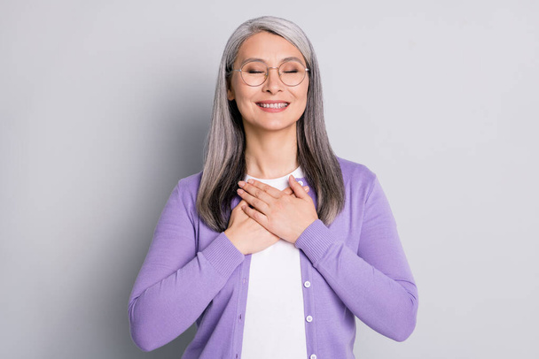 Photo portrait of elderly lady smiling holding two hands on chest with eyes closed wearing eyeglasses violet cardigan isolated on grey colored background - Foto, immagini
