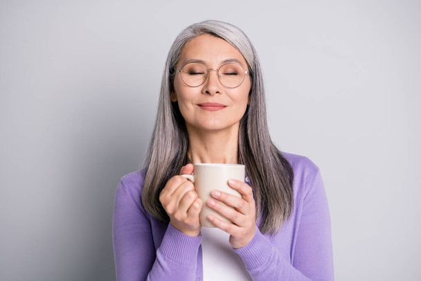 Photo portrait of woman holding cup of tea or coffee with two hands enjoying smell and taste with eyes closed wearing casual violet shirt isolated on grey colored background - Fotó, kép