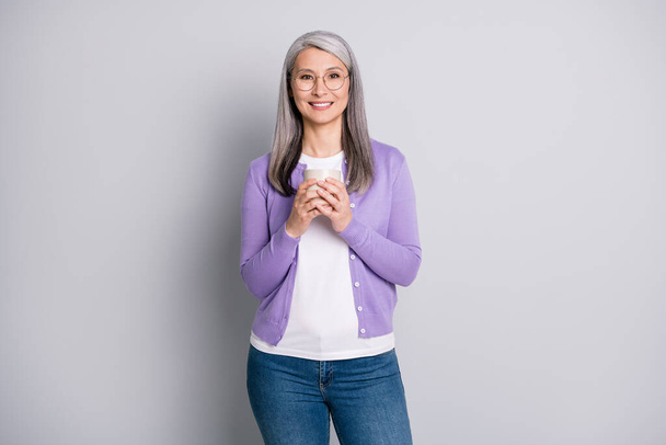 Photo portrait of old asian lady smiling holding cup of tea or coffee with both hands wearing violet cardigan isolated on grey colored background - Foto, afbeelding