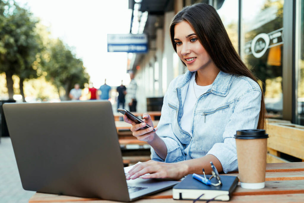 Young business woman working on open terrace at home, sitting in front of laptop, holding smartphone in her hand.  - Photo, image