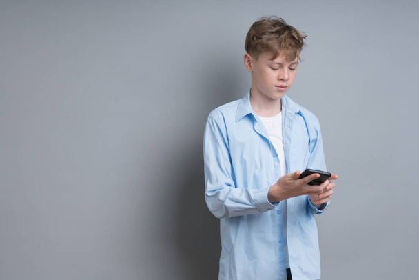 Egy kék inges, fehér pólós tinédzser szürke háttérrel játssza a telefont. Problémás tinédzser, szociális problémák, játékos, telefonfüggőség - Fotó, kép