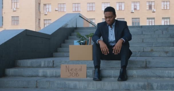 Desperacki afrykański biznesmen siedzi w pobliżu centrum biurowego z tabliczką Need Job, porażka biznesowa - Materiał filmowy, wideo